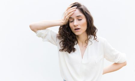 Exhausted tired woman with closed eyes touching head. Wavy haired young woman in casual shirt standing isolated over white background. Headache or burnout concept