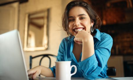 Cheerful attractive young woman enjoying distant work, sitting at desk using portable computer, drinking coffee. Pretty female blogger working from home, uploading video on her channel, smiling