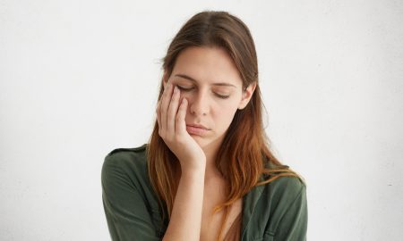 Cute woman having sleepy expression looking tired holding her hand on cheek closing her eyes with tiredness. Young woman having sad expression and toothache. People, problems, tiredness concept