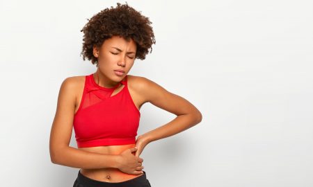 Dissatisfied African American woman holds aching hip, has kidney inflammation, touches location of pain near ribs marked with red dot, wears sport bra, shows perfect figure, has sporty shaped body