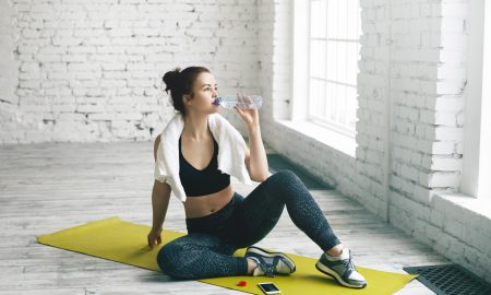 Health, sports, fitness, diet and weight loss concept. Beautiful young brunette female wiping sweat with towel after physical workout, sitting on mat and drinking fresh water out of plastic bottle