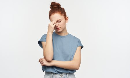 Portrait of tired, adult redhead girl with hair gathered in a bun. Wearing blue t-shirt and jeans. Touching bridge of her nose, suffer from headache. Stand isolated over white background