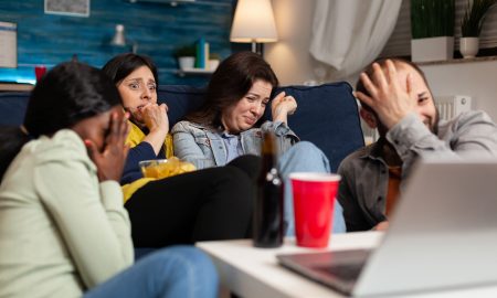 Multiracial friends chilling on sofa sitting in front of television watching horror thriller movie during night series. Group of multi-ethnic people having scary reaction, screaming with emotions.