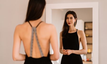 Woman in a long black dress with bare back looking at her reflection in the mirror while getting ready for an event, party, date