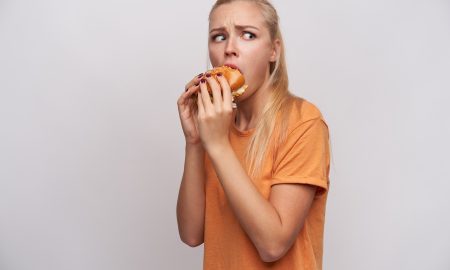 Portrait of confused young pretty blonde female with casual hairstyle looking thrillingly aside and rounding eyes while having lunch with fastfood, isolated over white background