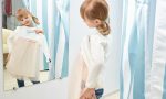 Reflection in mirror of cute girl. Little girl choosing and trying pink and white dress in fitting room with blue curtains. Child looking at mirror in mall or clothing store.