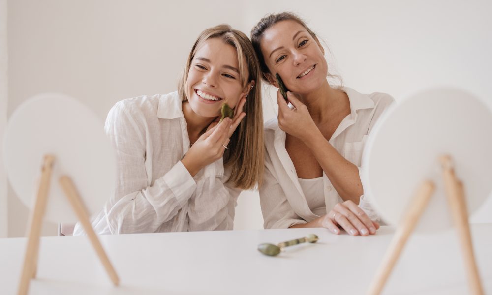 positive-young-caucasian-women-smiling-look-mirror-while-sitting-table-using-facial-scraper-medical-beauty-treatments