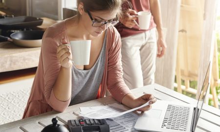 Young female drinking tea and studying bill in her hands, having frustrated look while managing family budget and doing paperwork, sitting at kitchen table with papers, calculator and laptop computer