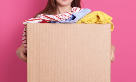 Indoor portrait of faceless girl standing with cardboard box in hands, holding carton box full of fashionable clothes isolated on rosy background. Donation, charity and volunteering concept.