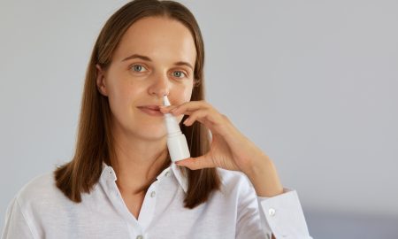Closeup indoor shot of charming woman using nasal spray for runny nose, catches cold, looking at camera, wearing white shirt, posing against light wall.